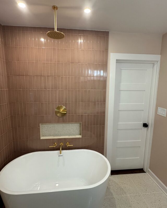 Pink stacked tile tub and shower wall with built-in niche, brass rain shower, and freestanding tub