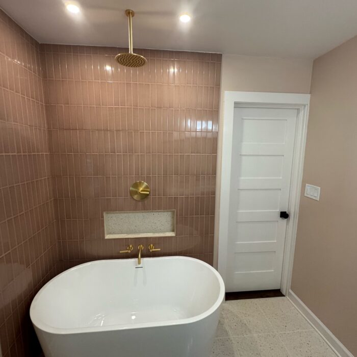 Pink stacked tile tub and shower wall with built-in niche, brass rain shower, and freestanding tub